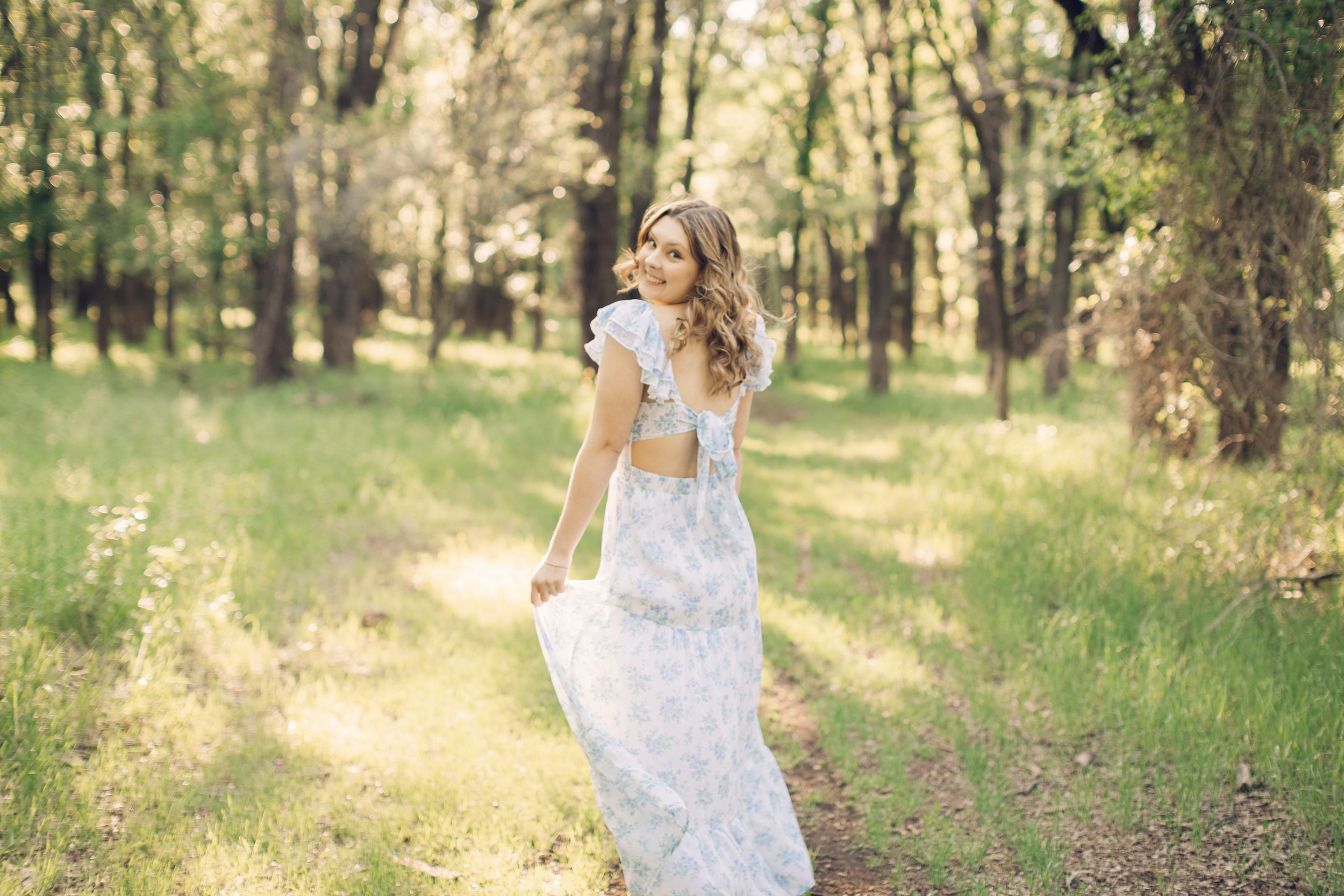 A senior girl in a floral blue and white dress twirls in a sunlit forest clearing.