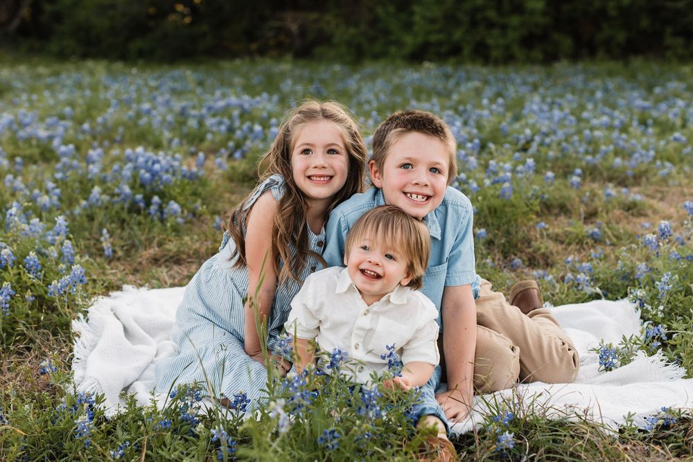 Three children sit together on a white blanket surrounded by blooming bluebonnets.