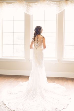 A bride in a flowing white wedding dress stands in a bright sunlit room with tall windows. Silouhette