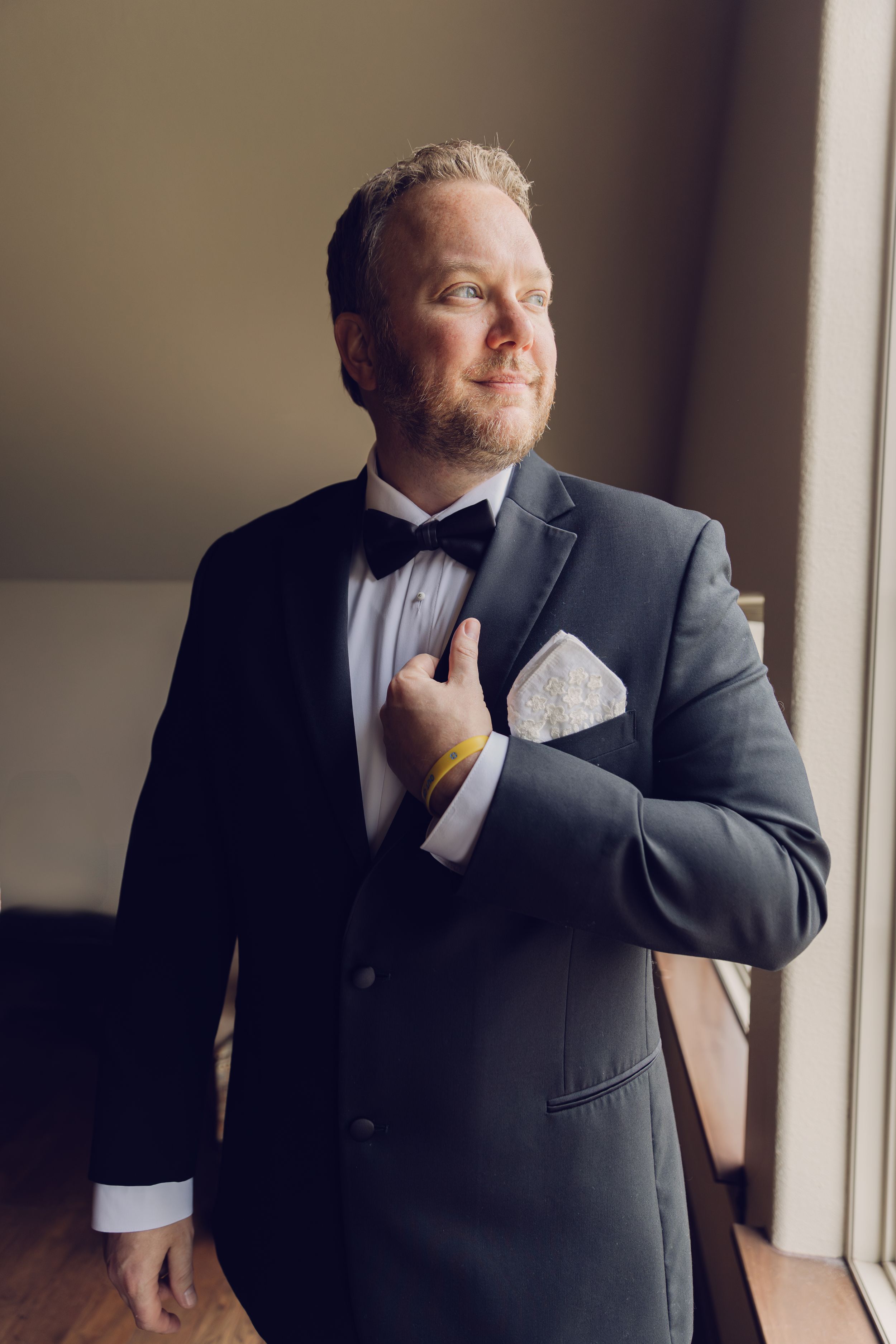 Groom in a dark suit adjusts his tie while standing by a window in natural light.
