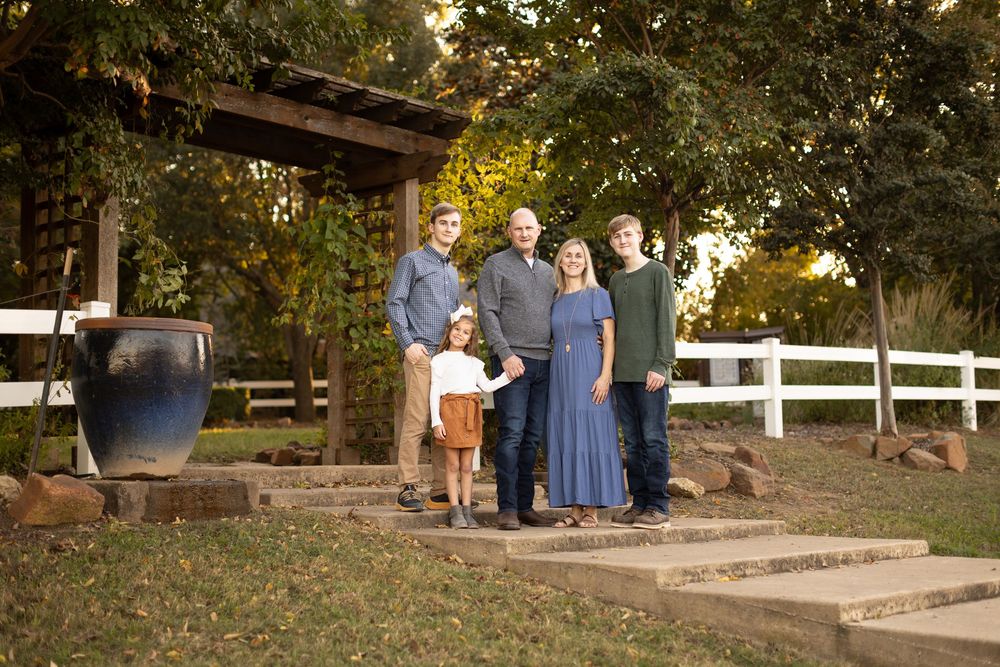 A family stands together on stone steps in front of a rustic building with white fencing.