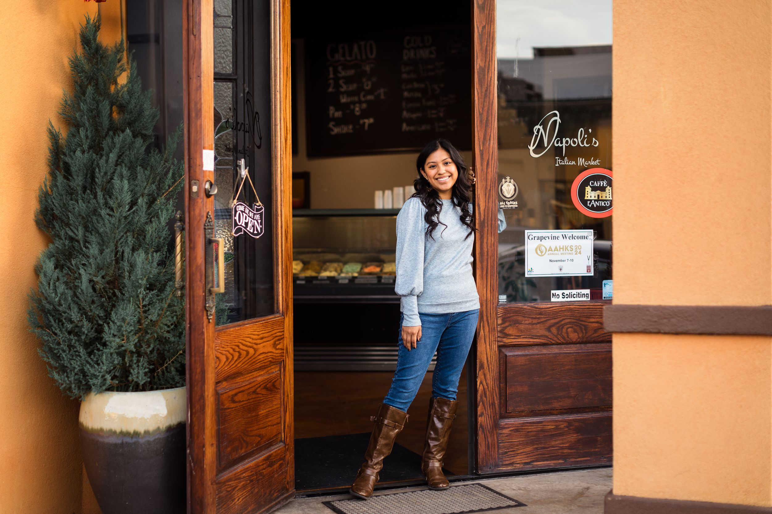 Senior girl in blue jeans and casual wear poses against a rustic brick storefront with wooden doors.