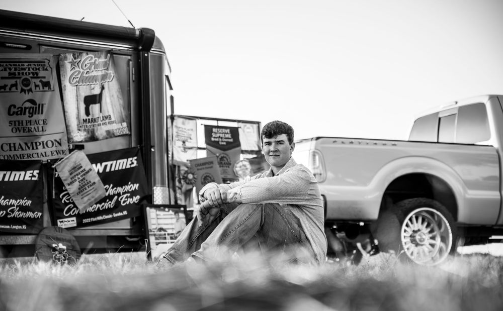 Senior boy sitting on the ground in front of a stock trailer with award banners from the fair.