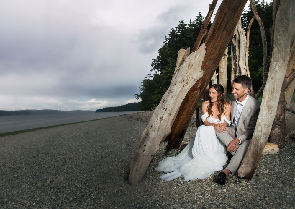 Bride and groom sitting together under driftwood on a sandy beach with evergreen trees and cloudy skies.
