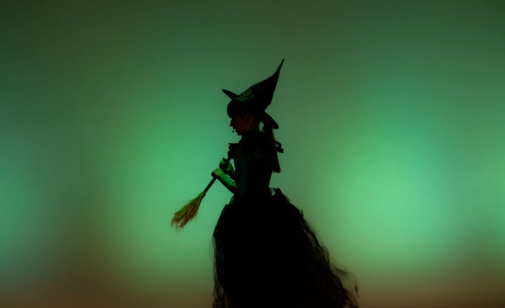 Silhouette of a witch in a pointed hat holding a broomstick against a green and teal glowing background.