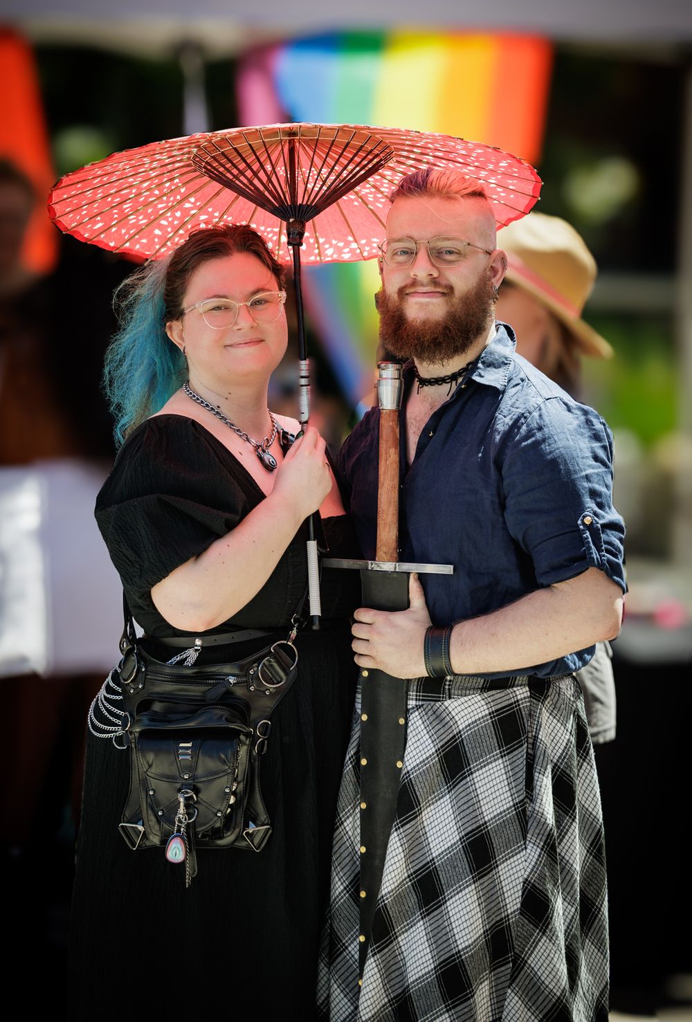 Two people smiling under a red parasol umbrella at a Pride event with rainbow flag in background.
