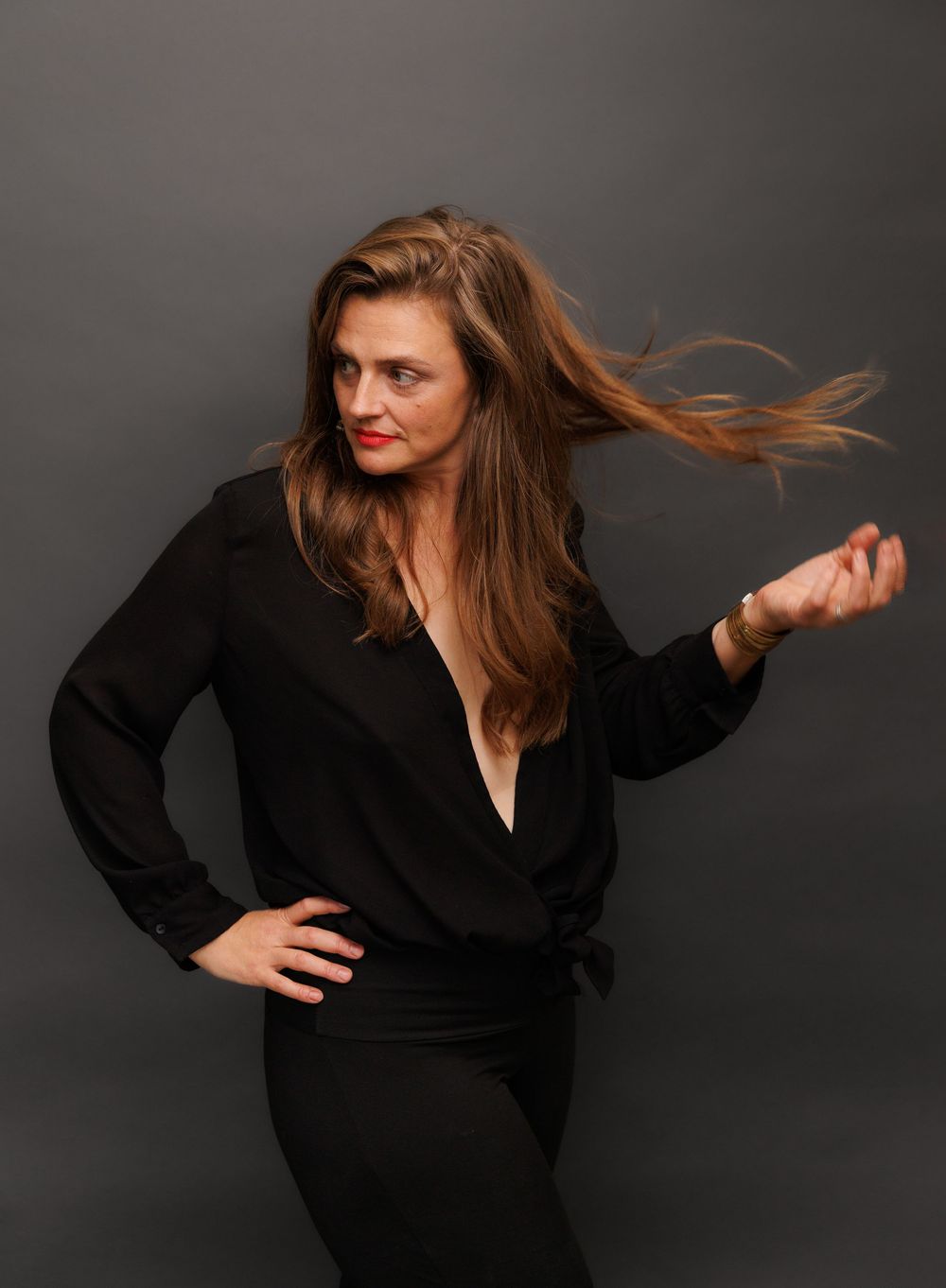 Woman in black wrap top with long flowing hair mid-motion against gray studio background.
