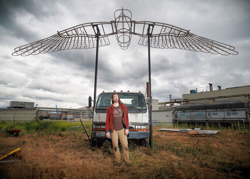 Person standing under large metal wing sculpture outdoors near industrial building.