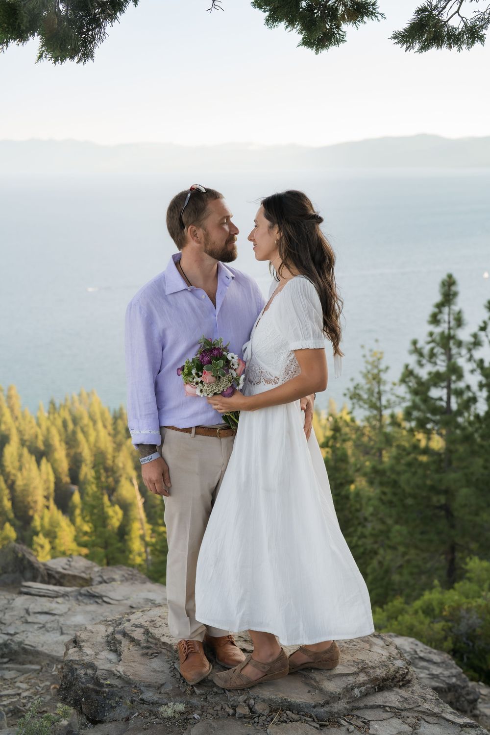 A couple sits together on a scenic mountain overlook during sunset, surrounded by evergreen trees and mountains.