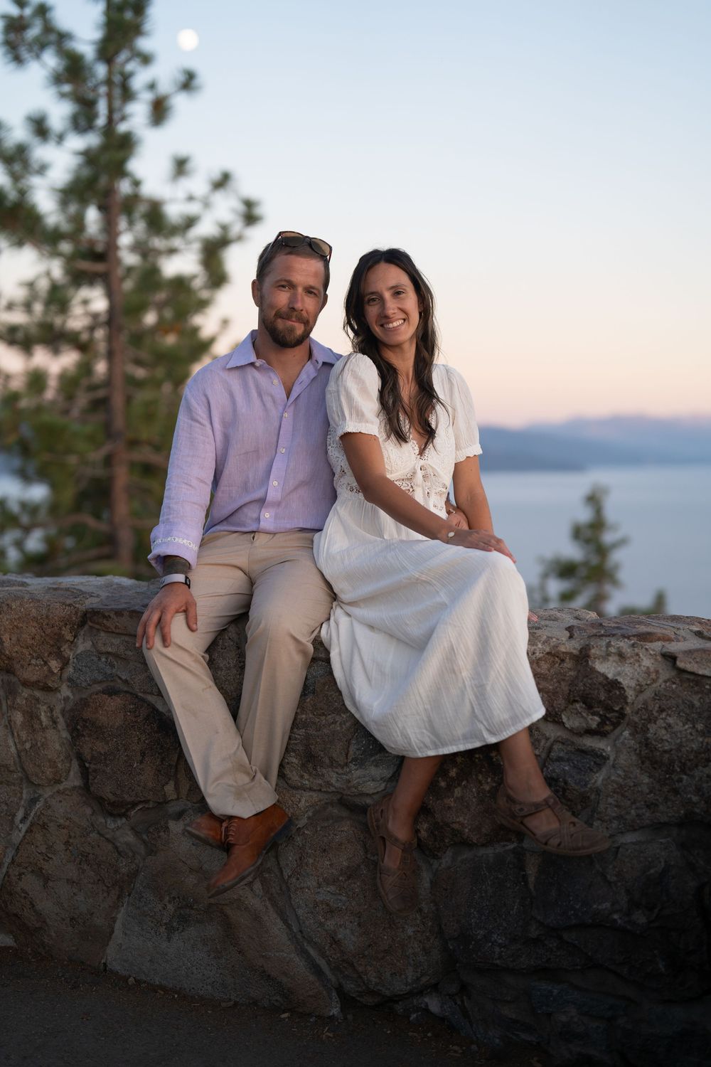 A couple sits together on rocky coastal outcrop at sunset with pine trees and water in background.