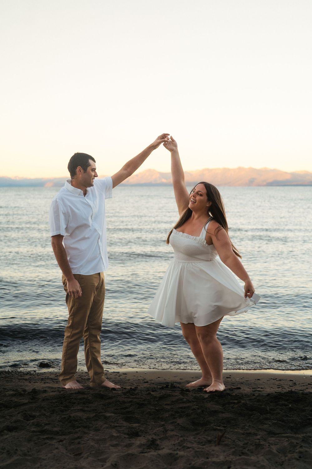 A couple dances together on a beach at sunset with mountains visible across the water.