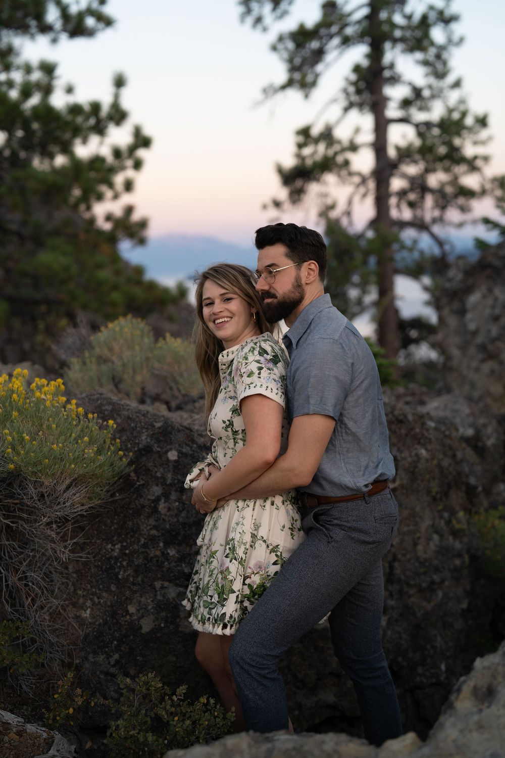A couple embraces on a rocky mountain overlook at sunset with pine trees and yellow wildflowers in the background.