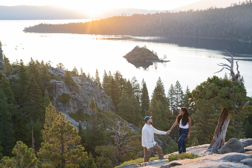 A scenic mountain viewpoint at sunset with evergreen trees and rocky cliffs overlooking a serene lake.