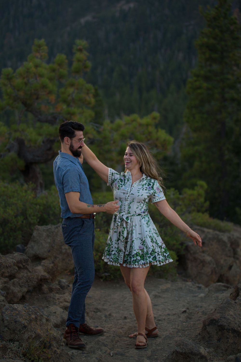 A couple shares a playful moment while posing together on a rocky overlook with evergreen trees in the background.
