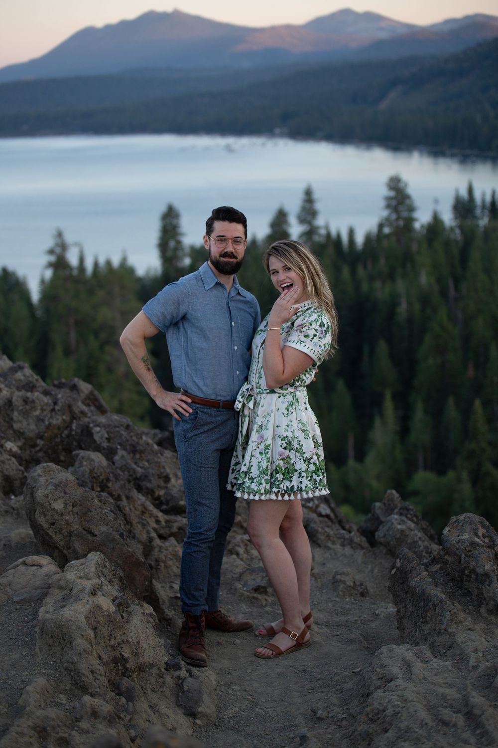 A couple poses together at sunset overlooking Lake Tahoe with mountains and forest in the background.