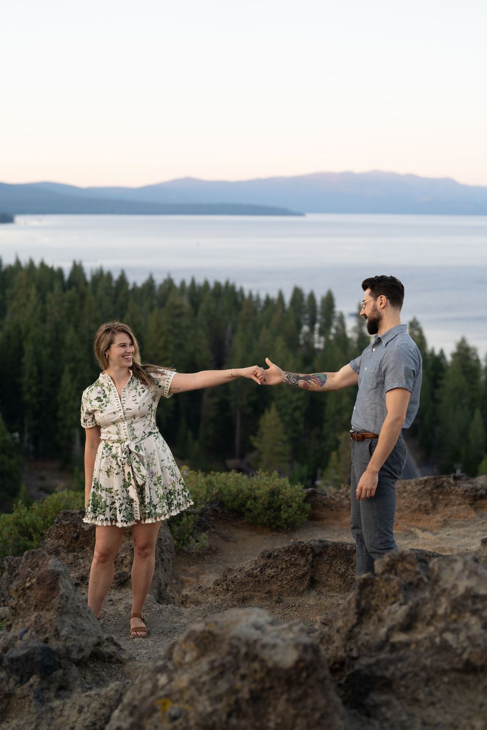 A couple stands on a rocky mountain overlook with Lake Tahoe and mountains visible in the background at sunset.