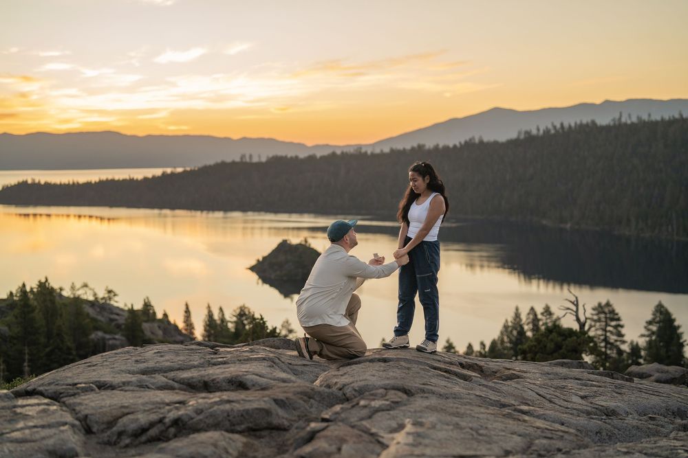 Two people stand on a rocky overlook at Emerald Bay Lake Tahoe during a golden sunset with mountains in the distance.
