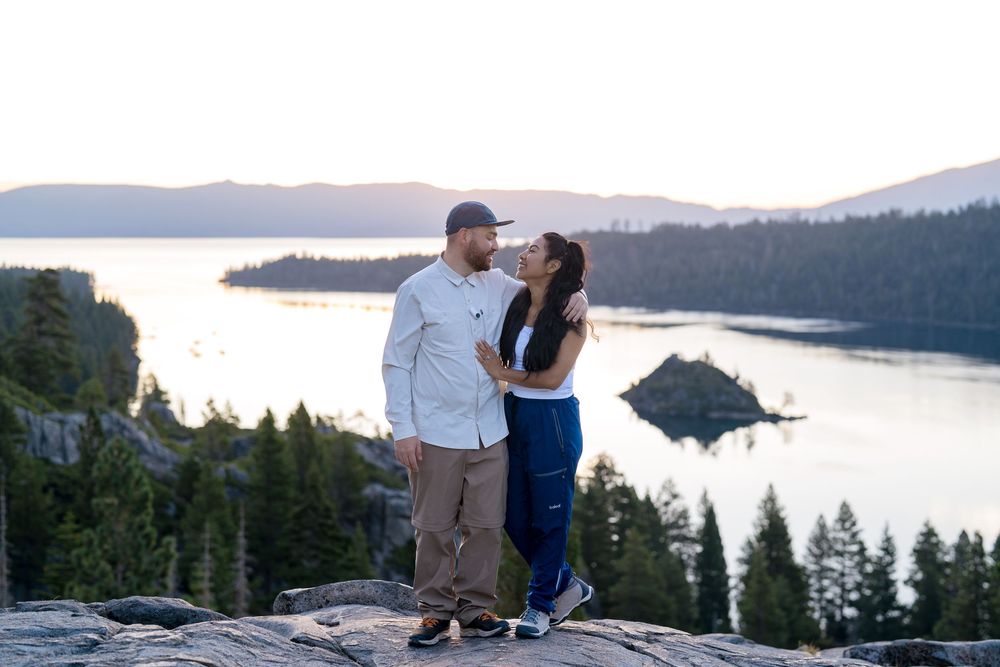 A couple stands together on a cliff overlooking Emerald Bay and Fannette Island at Lake Tahoe during dusk.