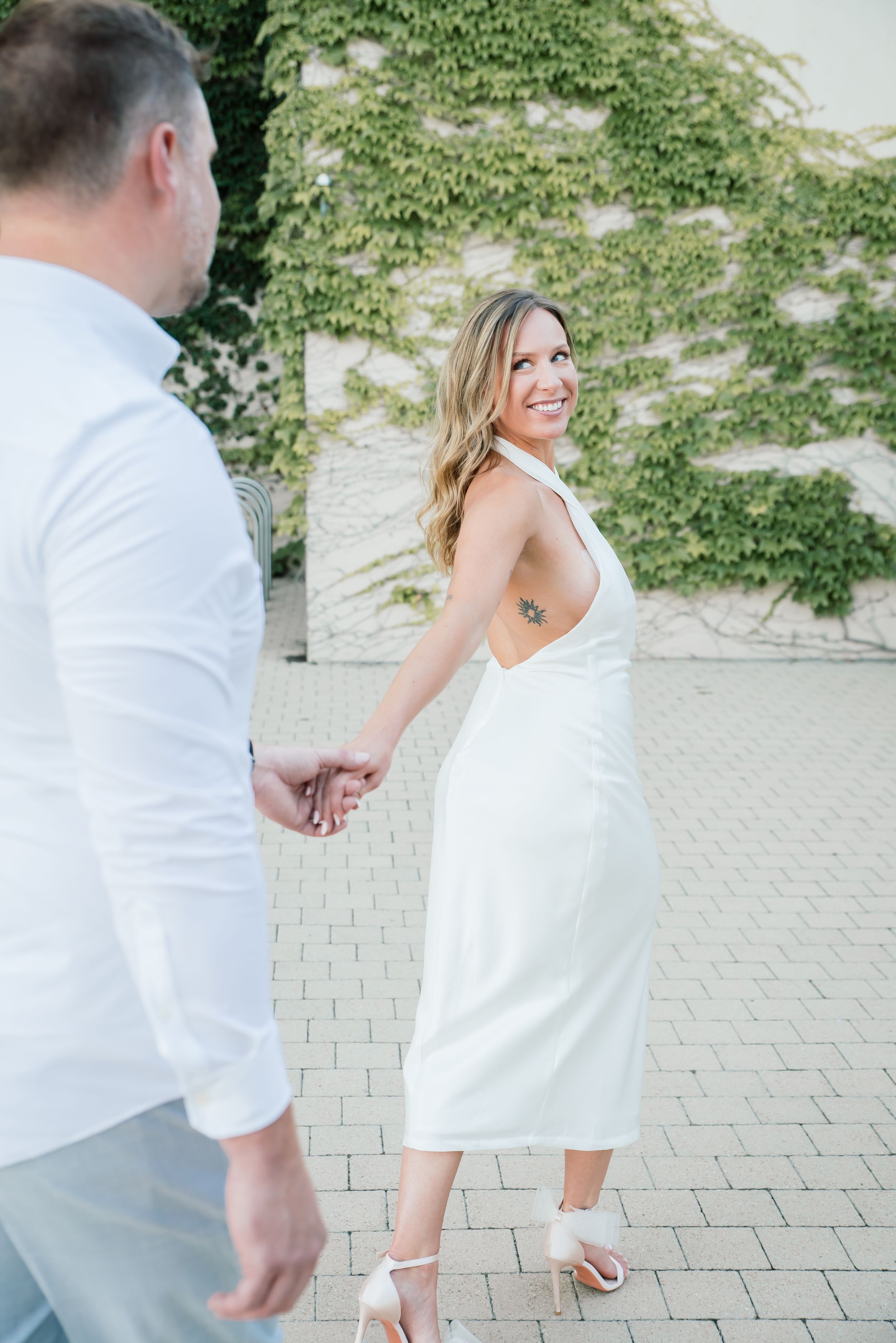 Smiling bride in white backless dress holds hands with groom outdoors near ivy-covered wall.