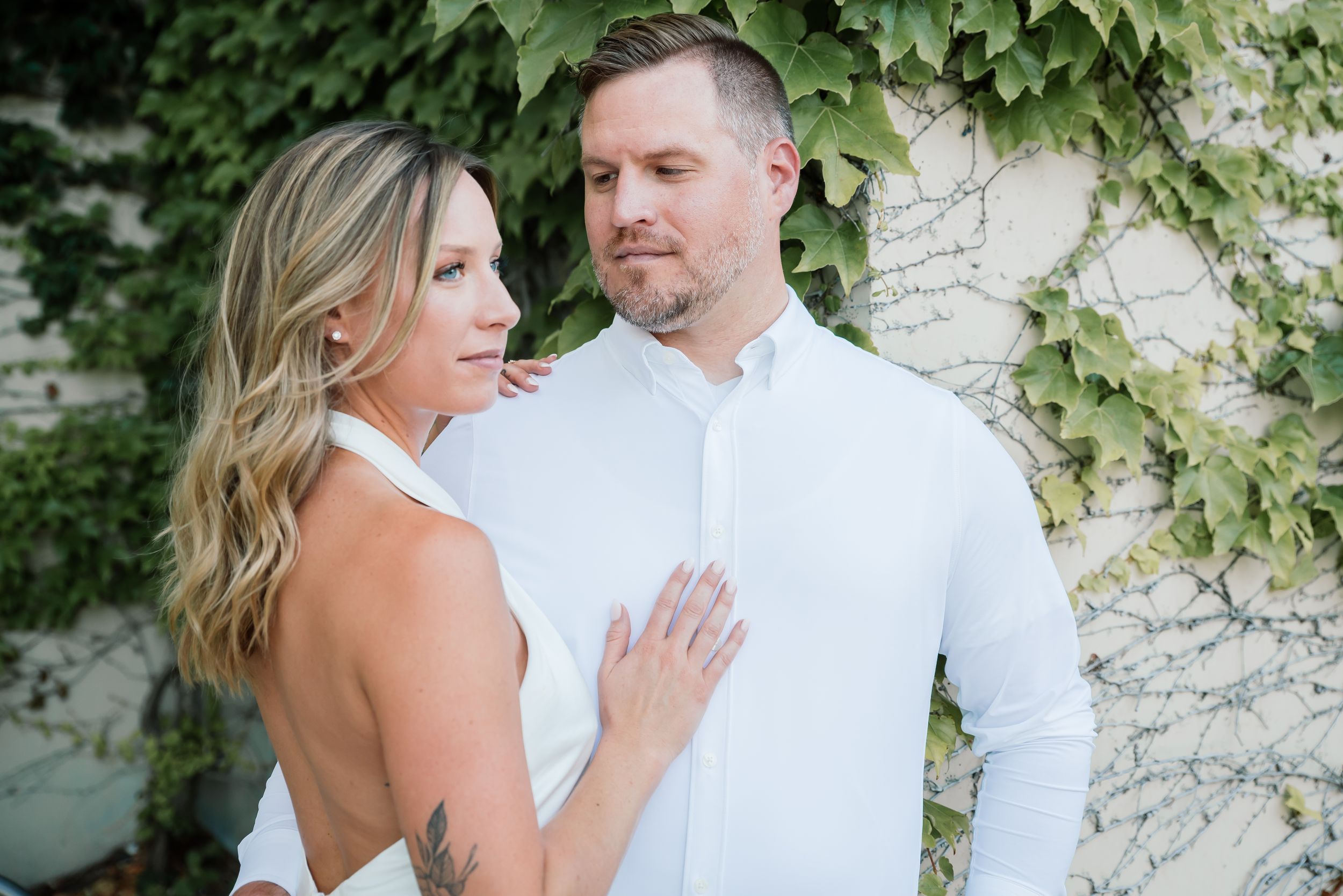 Engaged couple in white attire posing together against an ivy-covered wall outdoors.