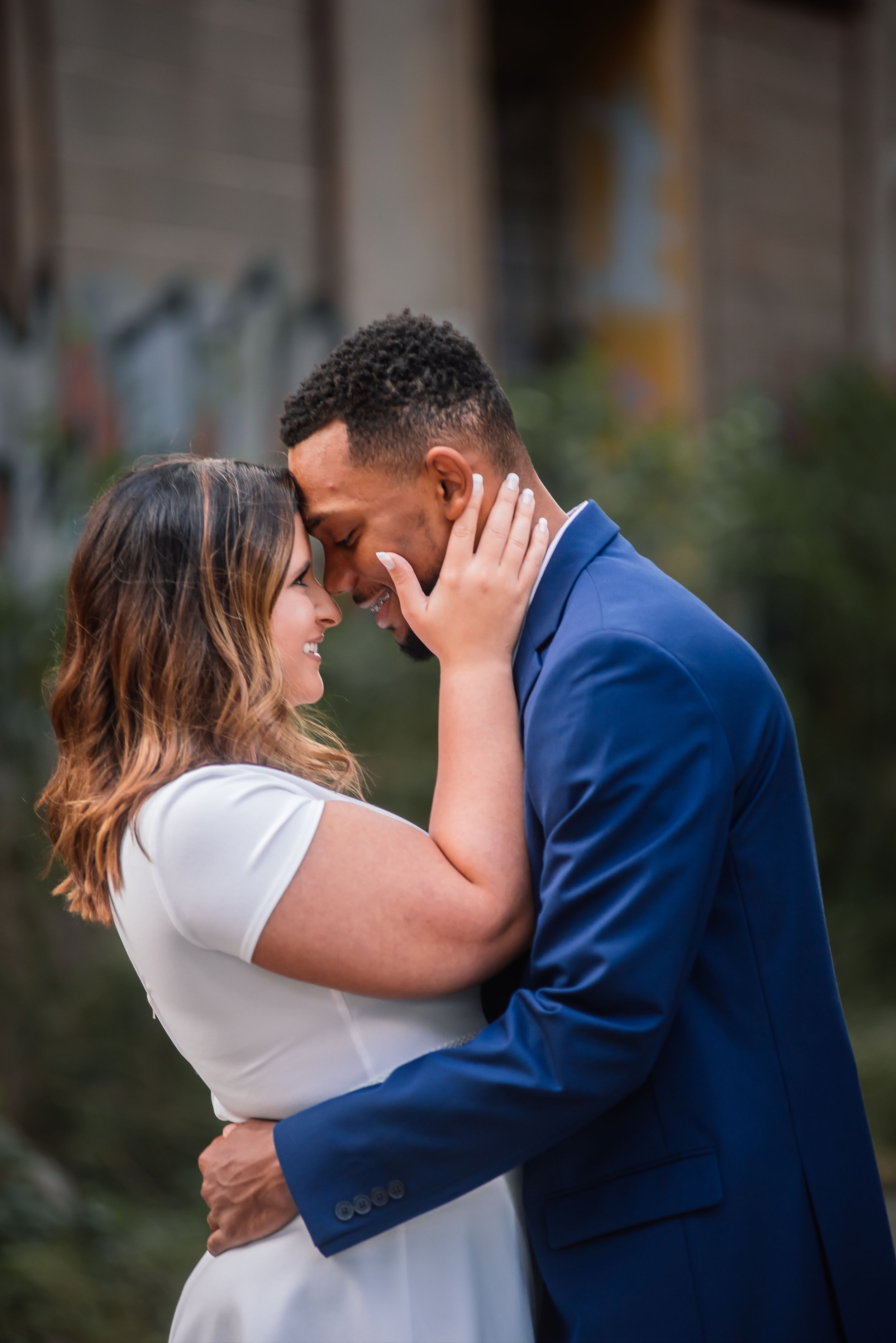 Engaged couple in a tender forehead-to-forehead embrace, man in blue suit, outdoors.