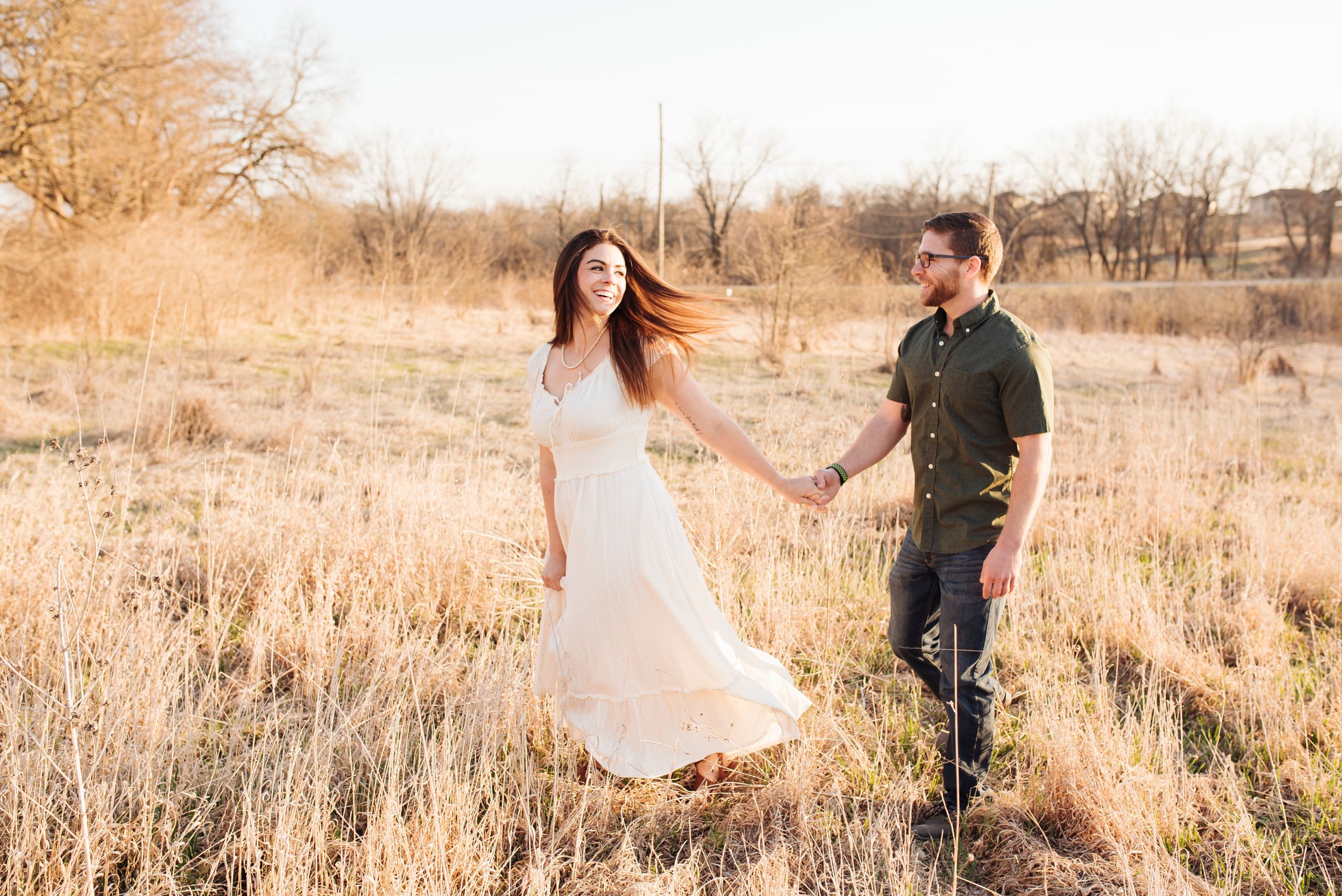 Woman in white dress holding hands with man while walking in golden field.