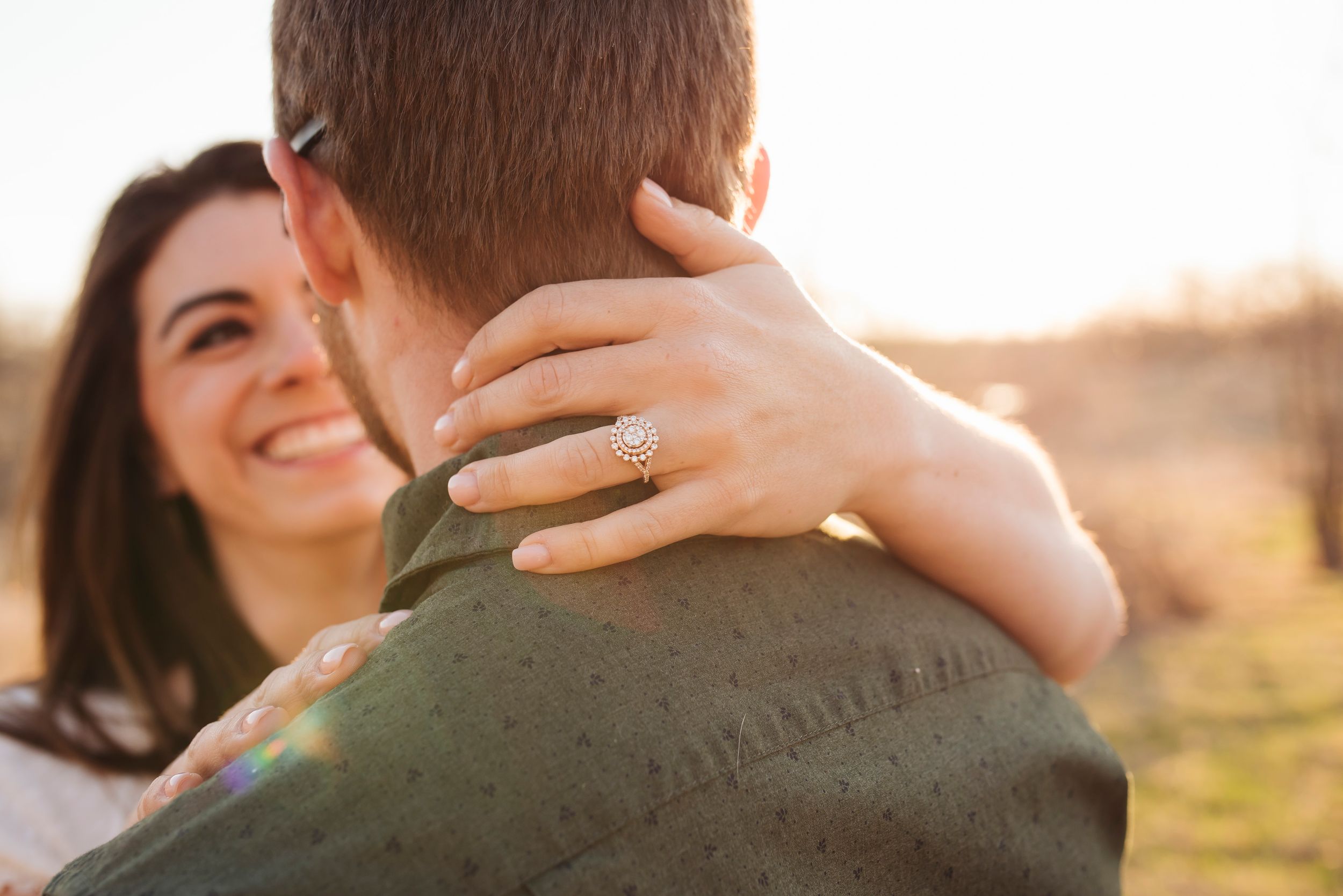 Smiling woman with engagement ring embracing man in golden sunset light.