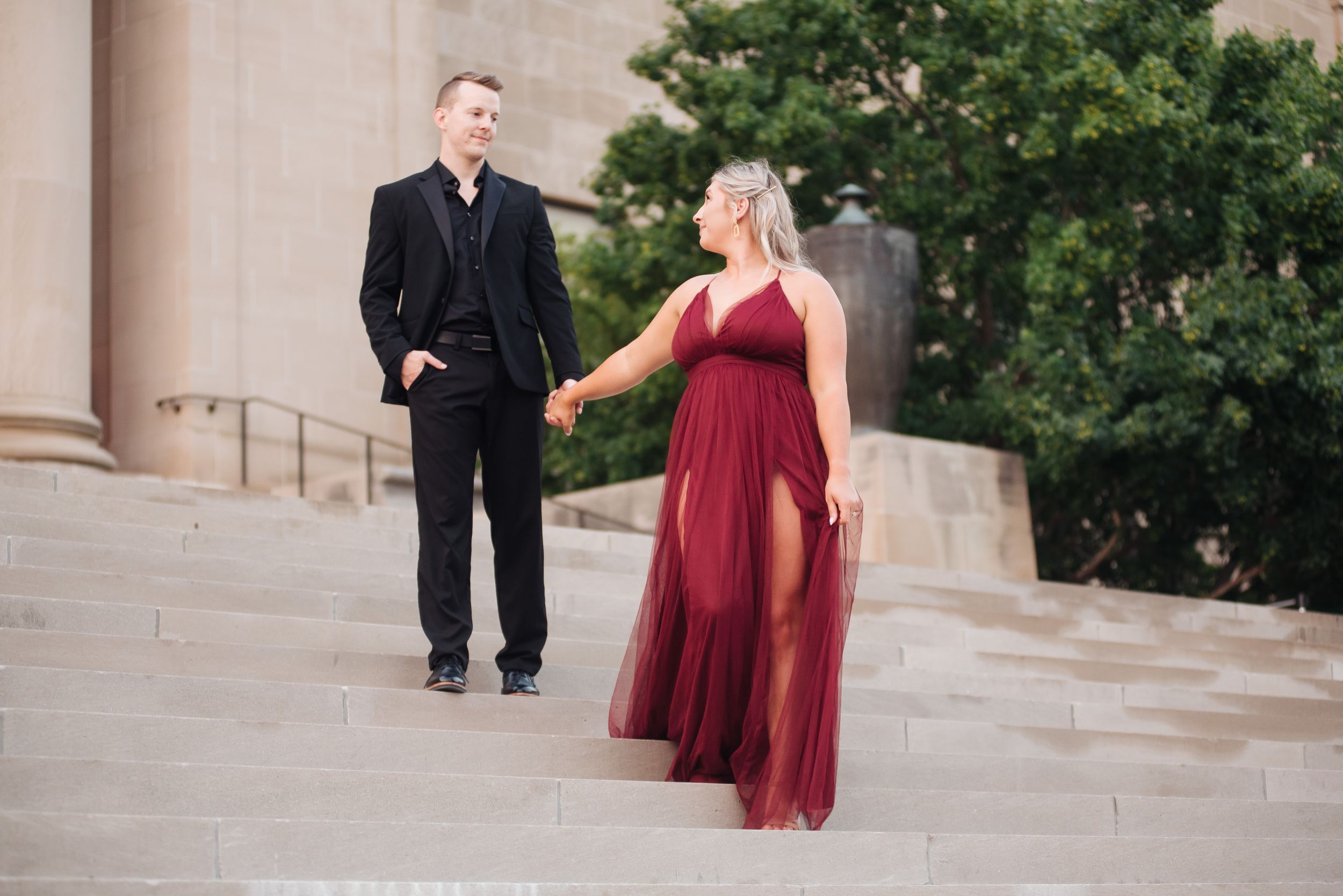Couple walking down grand stone steps, woman in flowing burgundy dress holding hands.