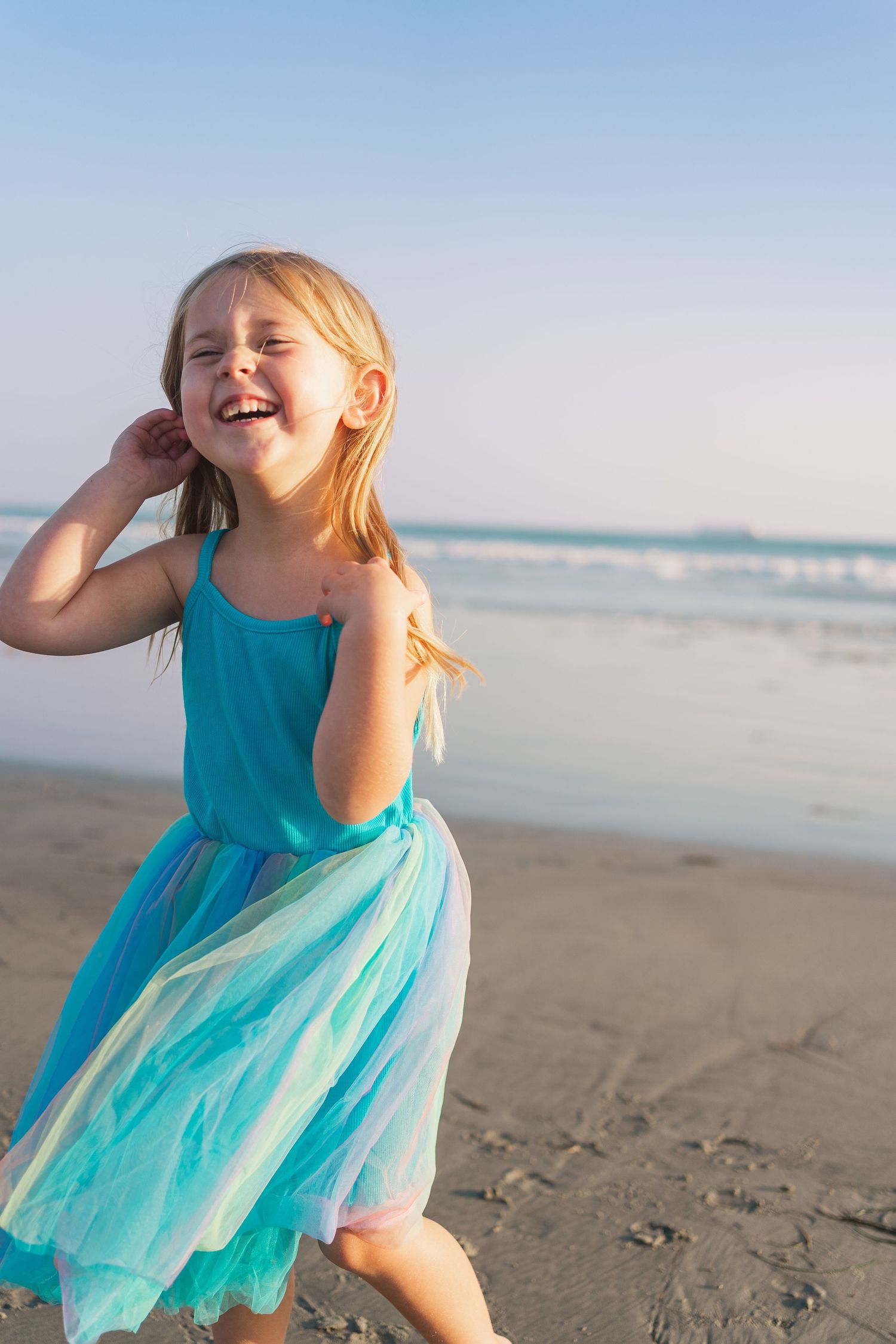 A child wearing a turquoise dress dances joyfully on a sunny beach with ocean waves in the background.