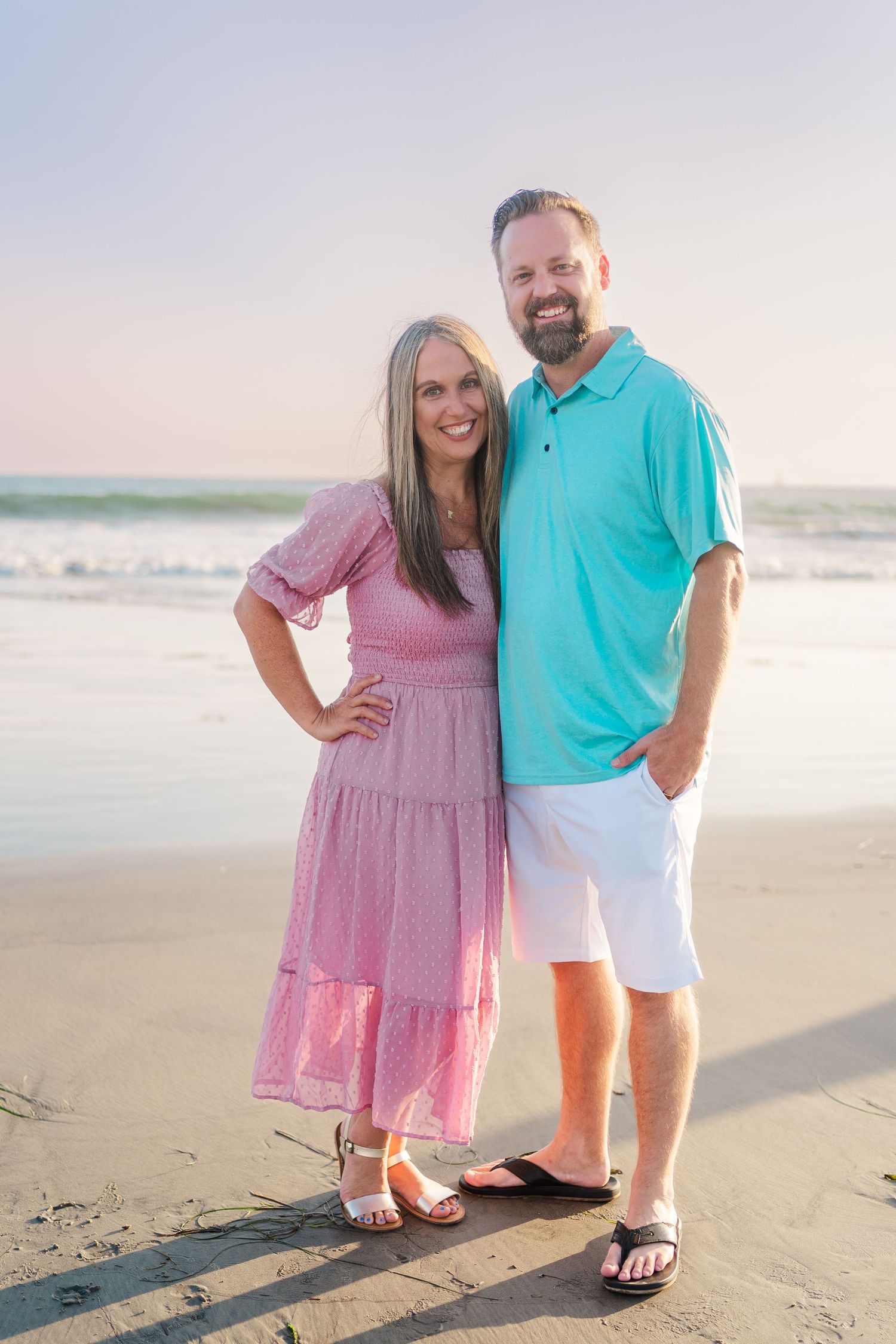 A couple stands together on a beach at sunset, wearing summer attire with a pink dress and turquoise shirt.