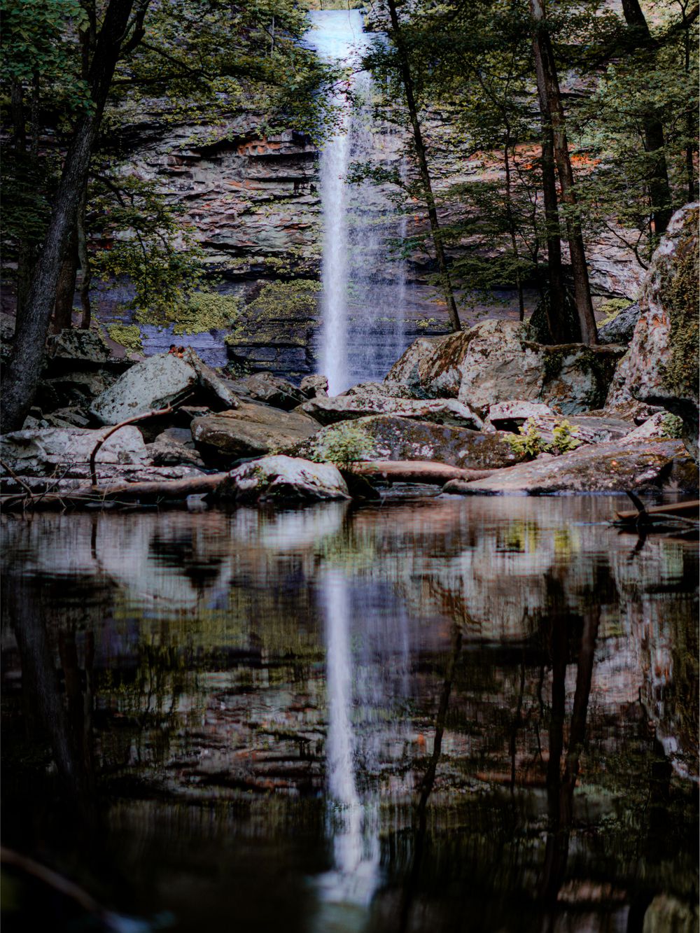 Tall waterfall cascades into a still reflecting pool surrounded by forest and rocky terrain.