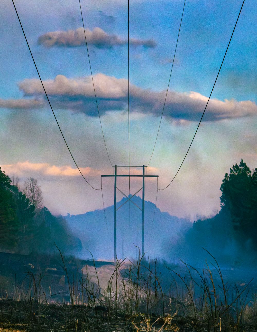 Power lines stretch toward a misty mountain silhouette under a cloudy blue sky.