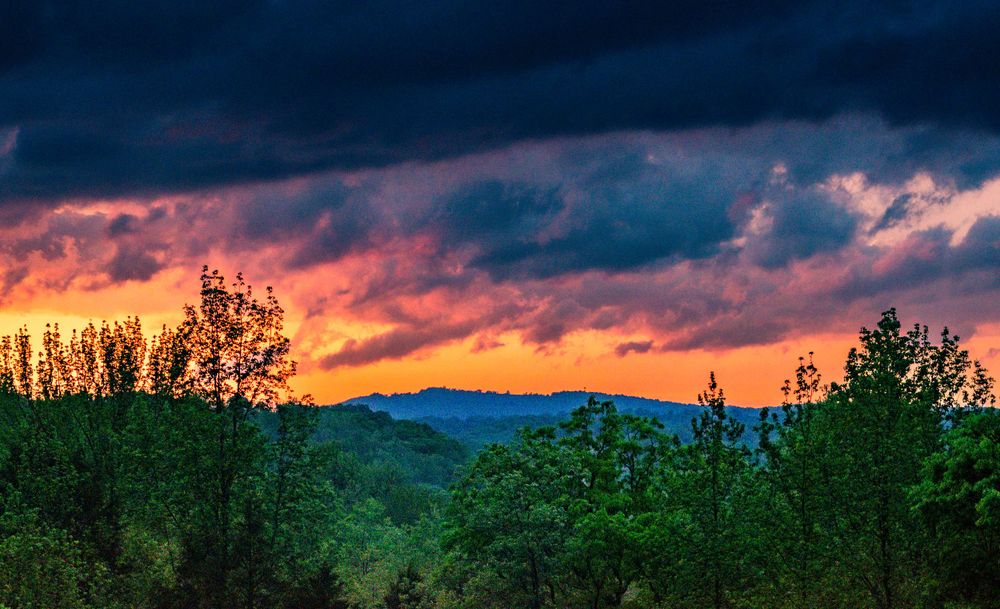 Dramatic sunset sky with dark storm clouds over mountain peaks and evergreen forest.