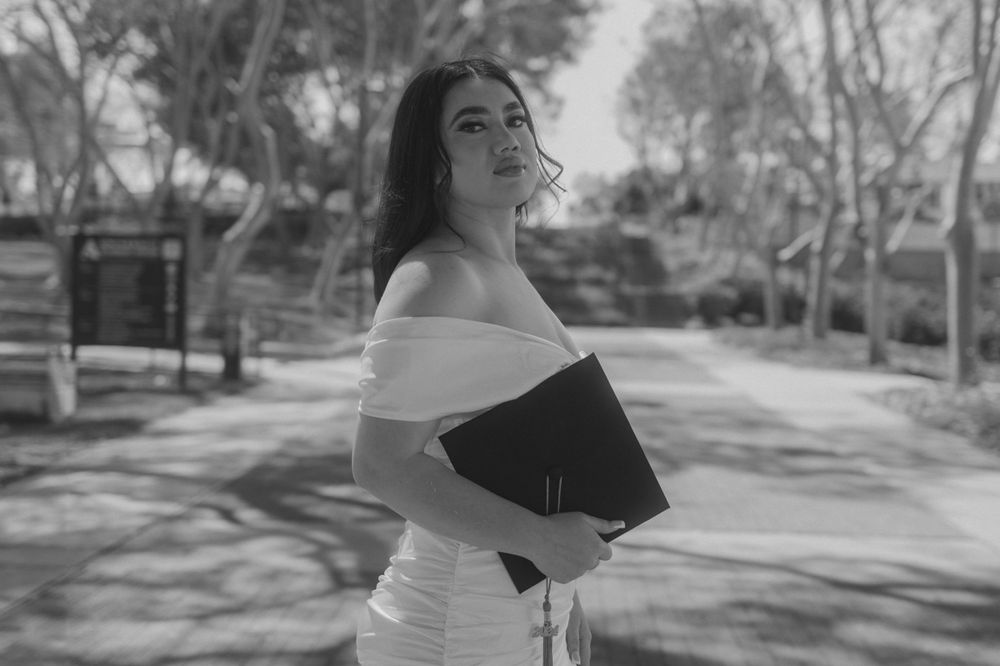 Woman in white dress holding black folder on tree-lined path.