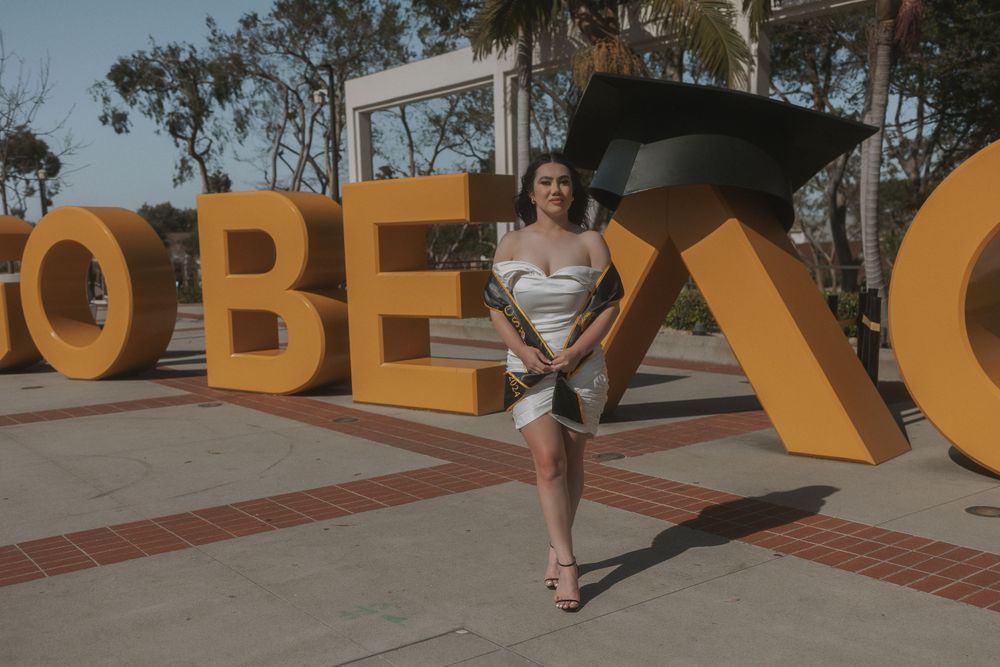 Graduate in white dress posing with large orange decorative letters in background.