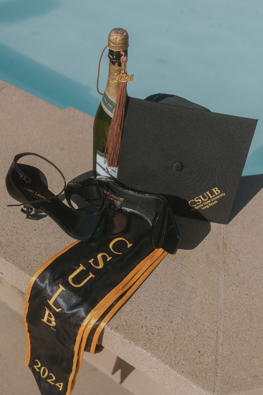Still life of graduation accessories including cap, stole, heels and champagne by pool.