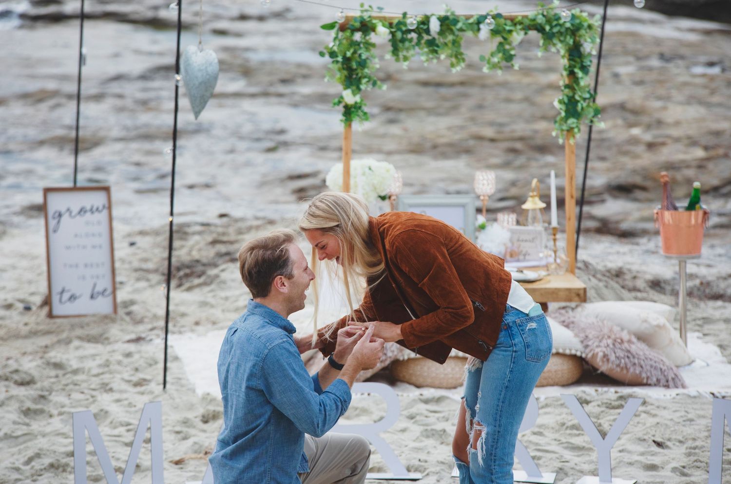 Beach proposal scene with decorated arch and flowers along the shore.