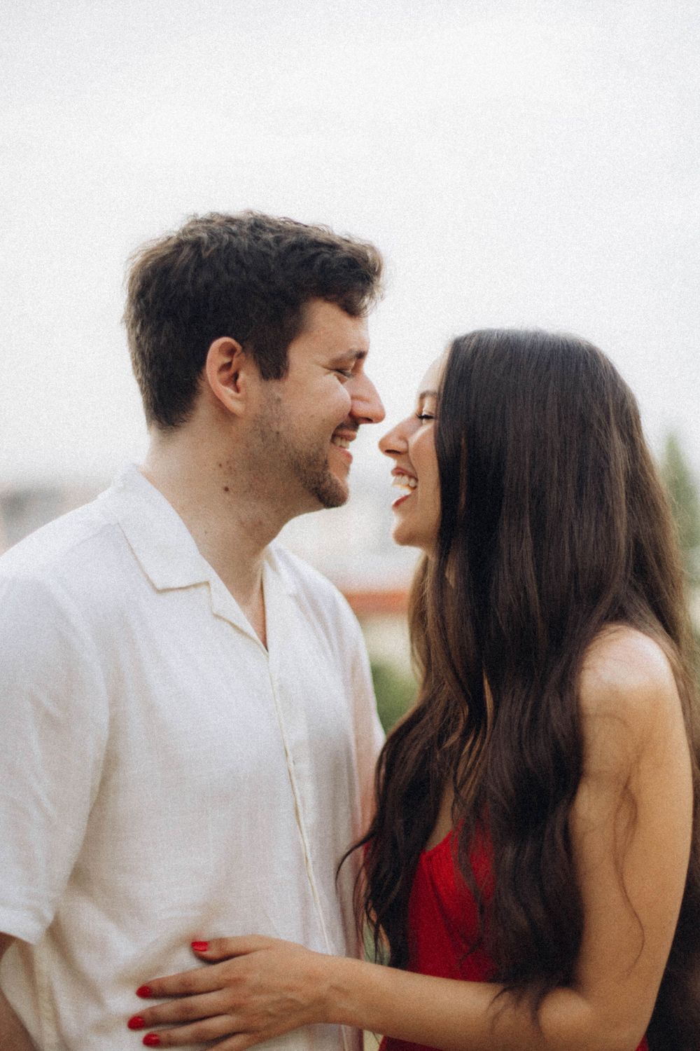 Romantic couple smiling face to face outdoors, woman in red dress with long dark hair, man in white shirt.