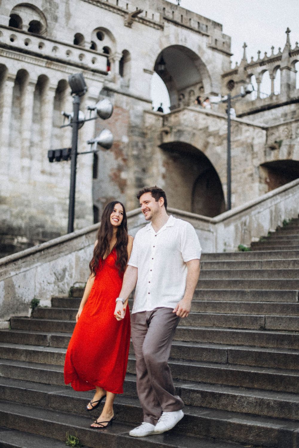 Couple holding hands walking down historic stone steps, woman in red dress, man in white shirt.