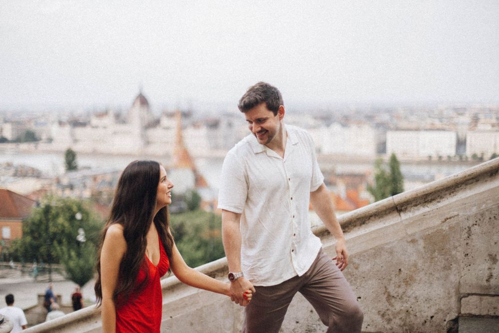 Smiling couple holding hands on stone steps with panoramic city skyline visible in the background.
