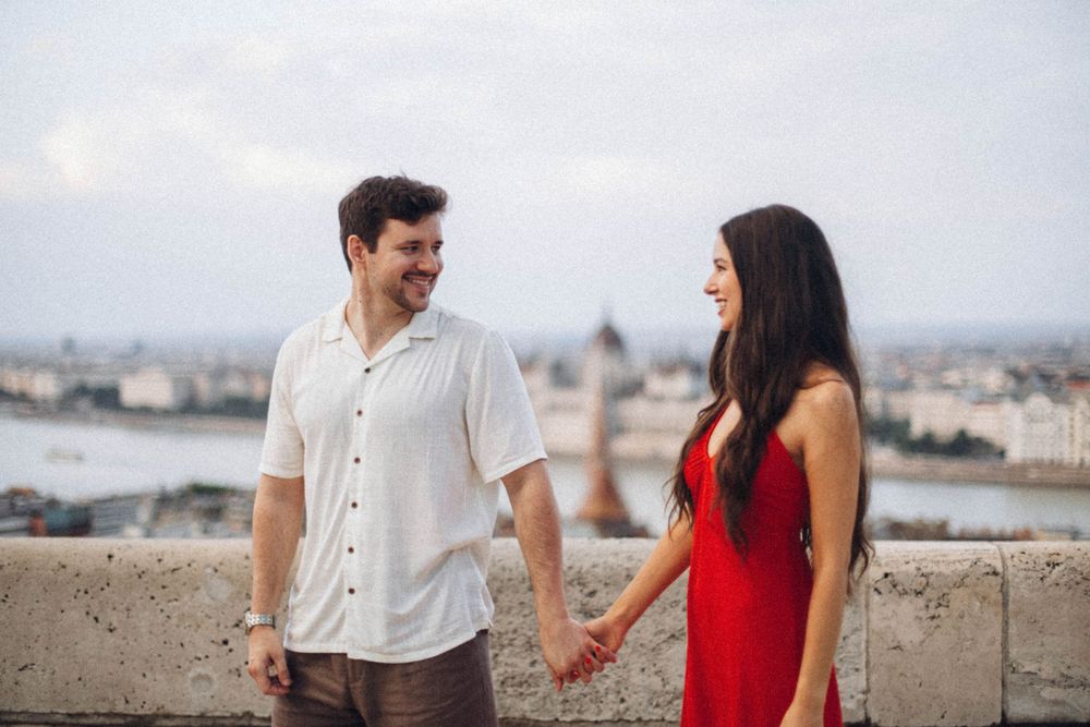 A smiling couple holds hands on a Budapest terrace with the Parliament building in the background.