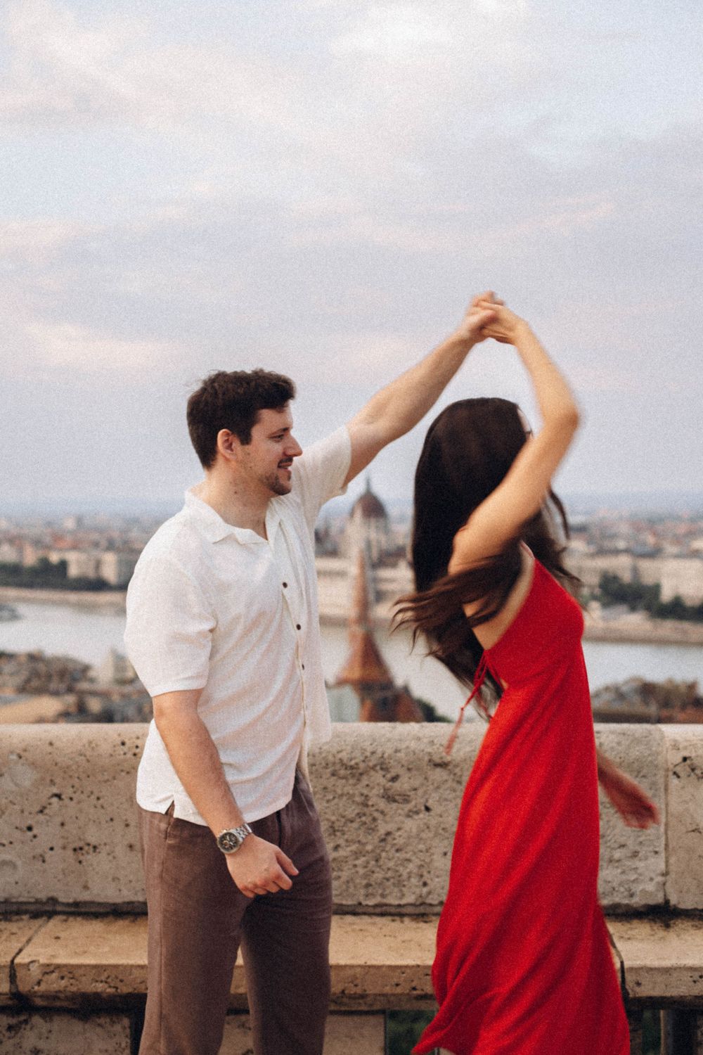 A laughing couple dances on a Budapest overlook with the iconic Parliament building visible in the distance.