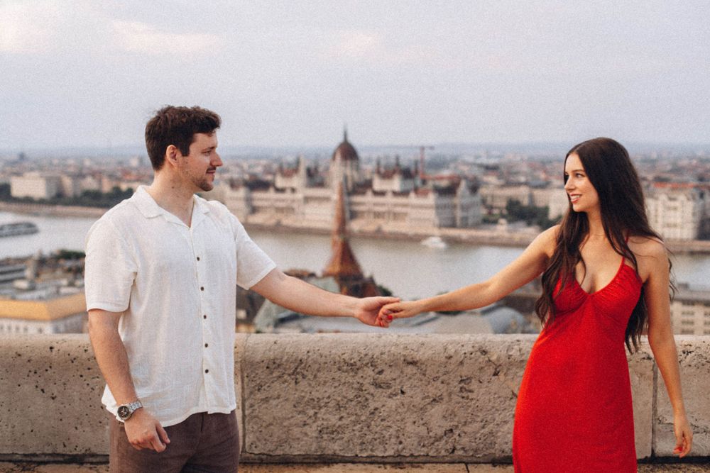 A romantic couple holds hands and smiles at each other on a Budapest terrace overlooking the Parliament.