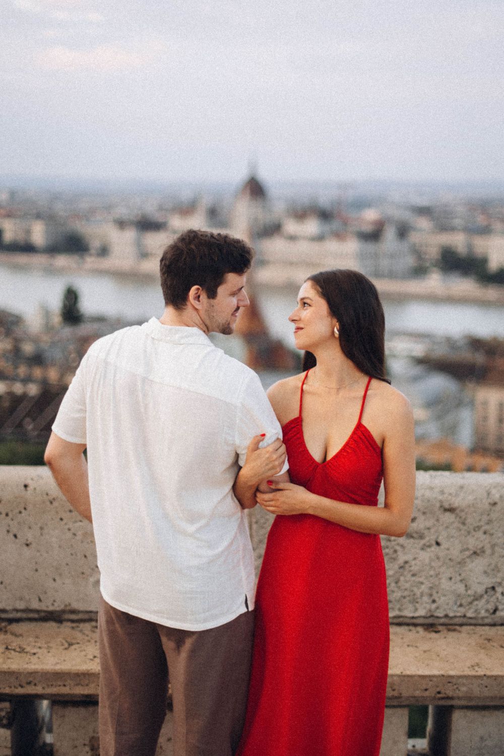 A couple embraces and gazes at each other on a Budapest terrace with the Parliament building behind them.