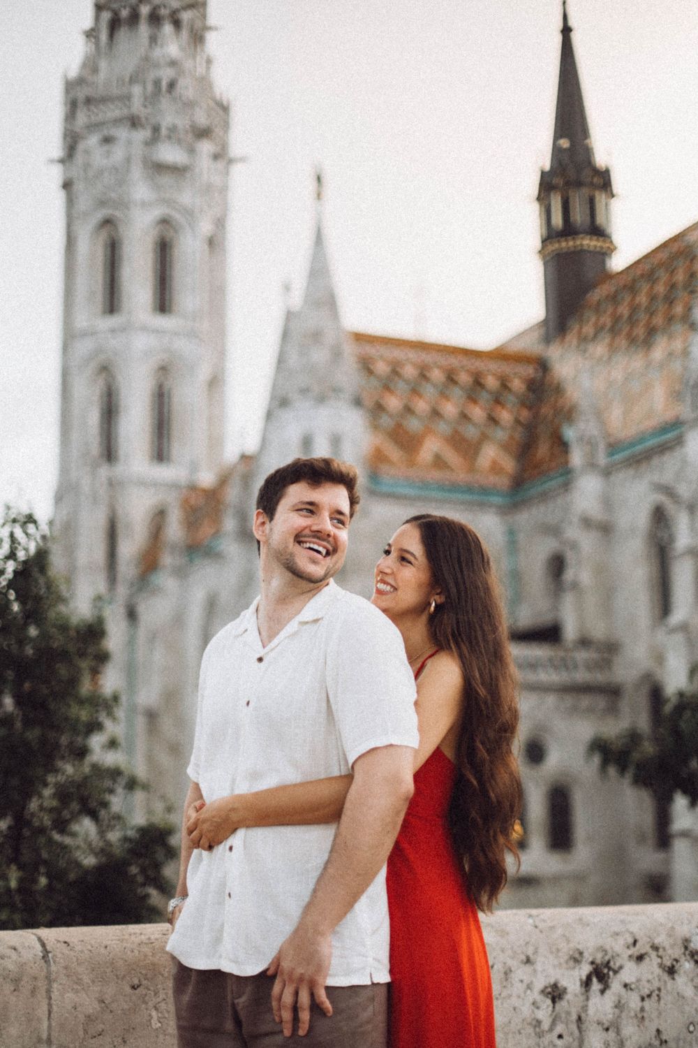 A smiling couple embraces in front of the historic Matthias Church in Budapest on a cloudy day.
