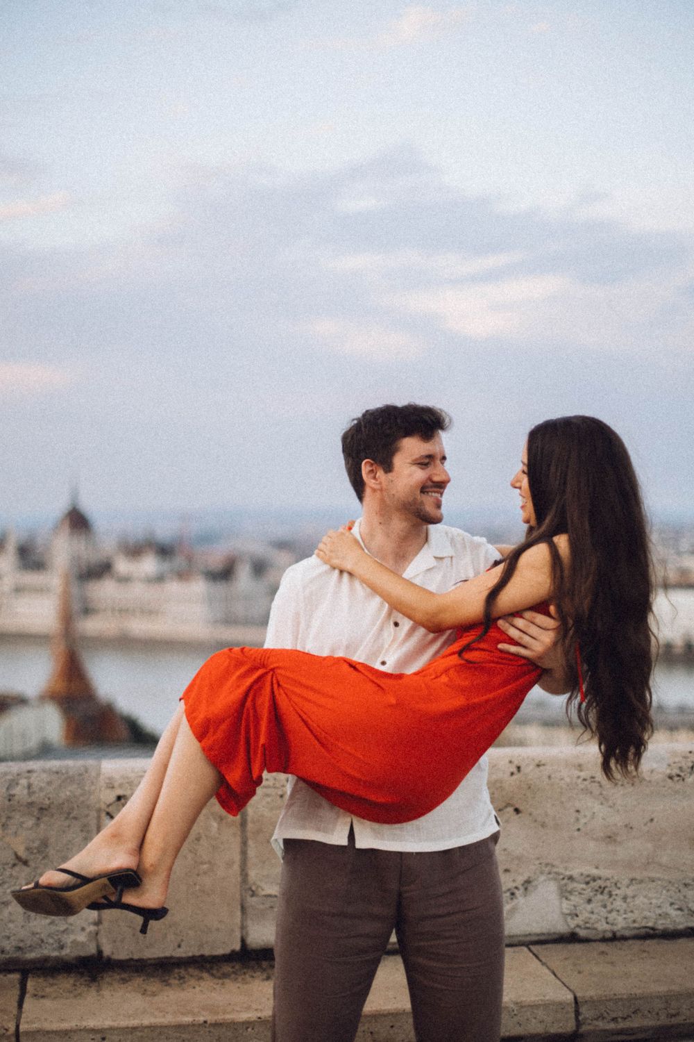 A man carries a woman in a red dress while smiling on a Budapest terrace with the Parliament behind them.