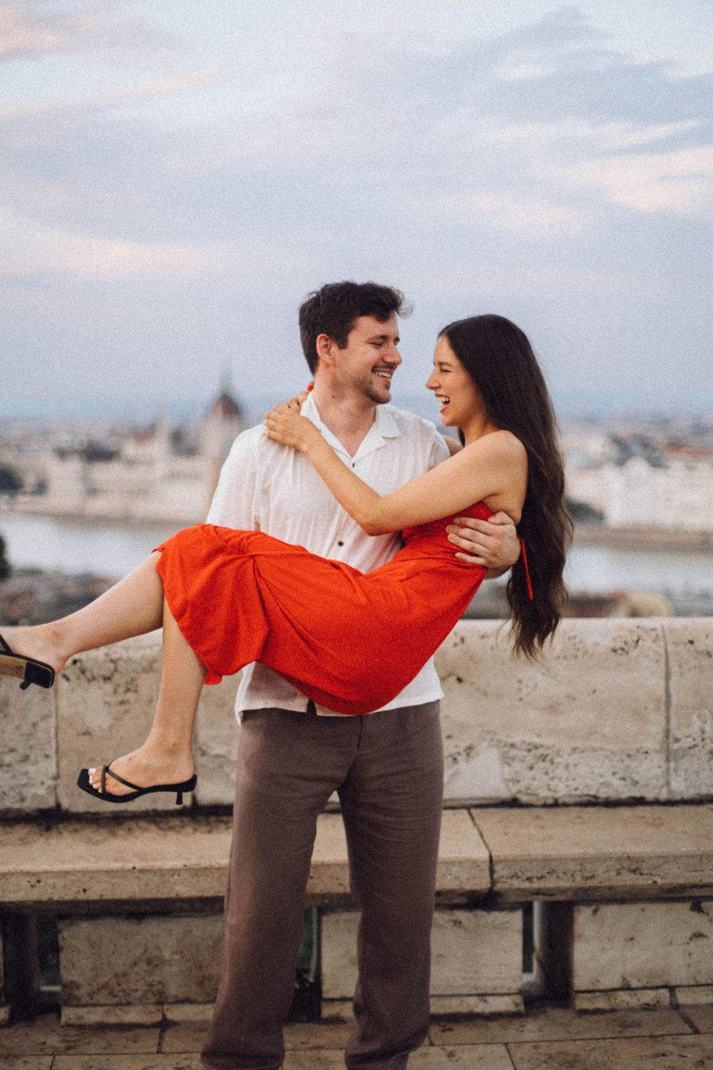 A laughing couple shares a joyful moment as the man carries the woman on a scenic Budapest overlook.