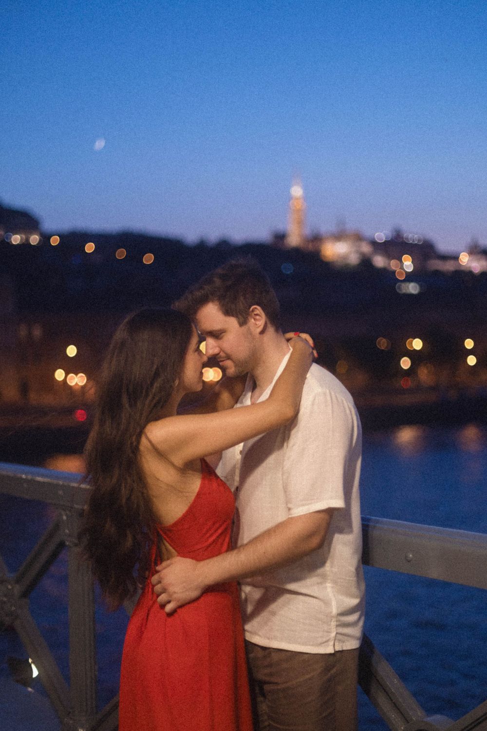 Romantic couple embracing on a bridge at night with Budapest city lights behind them.