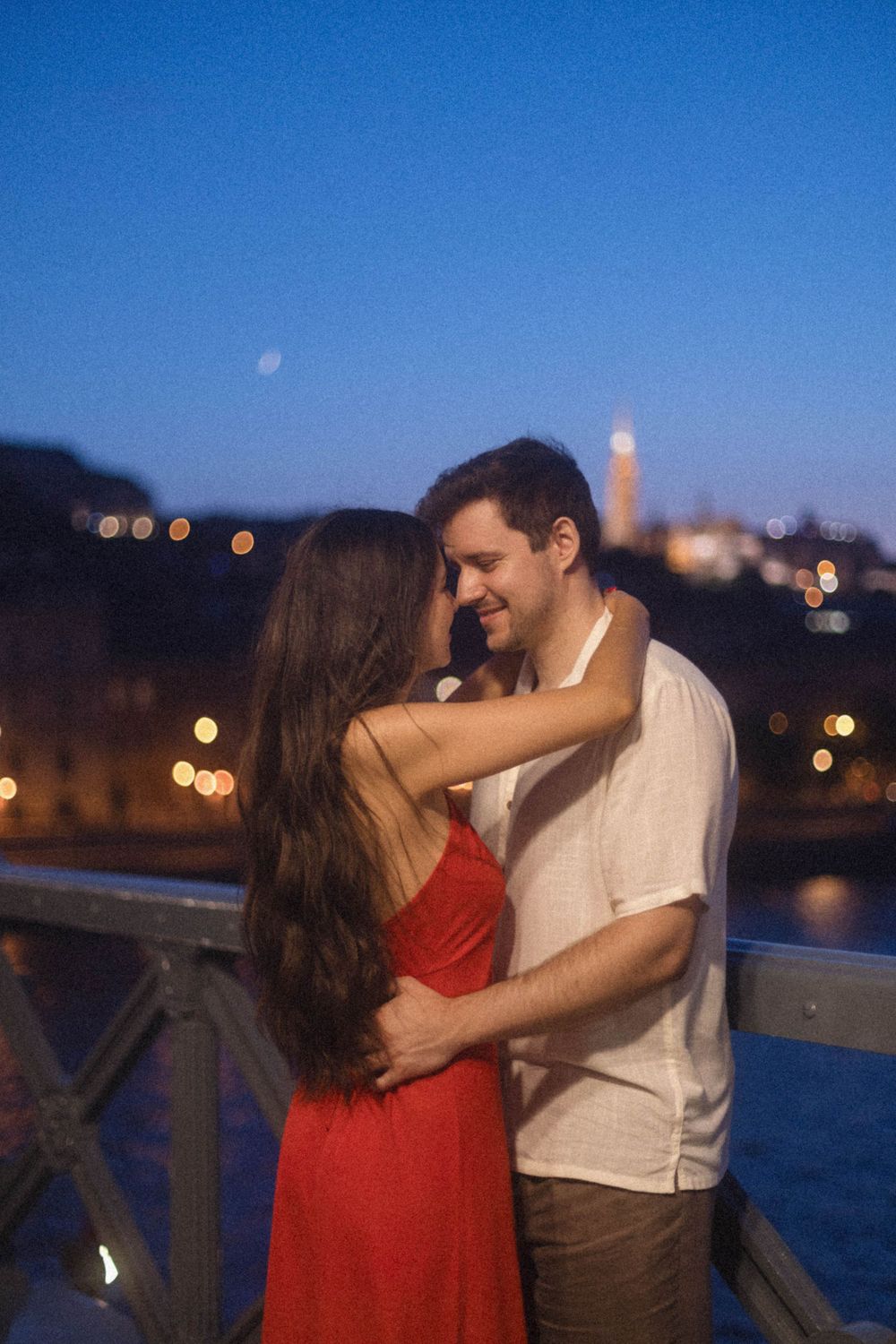 Couple in red dress and white shirt sharing an intimate moment on a bridge at dusk.