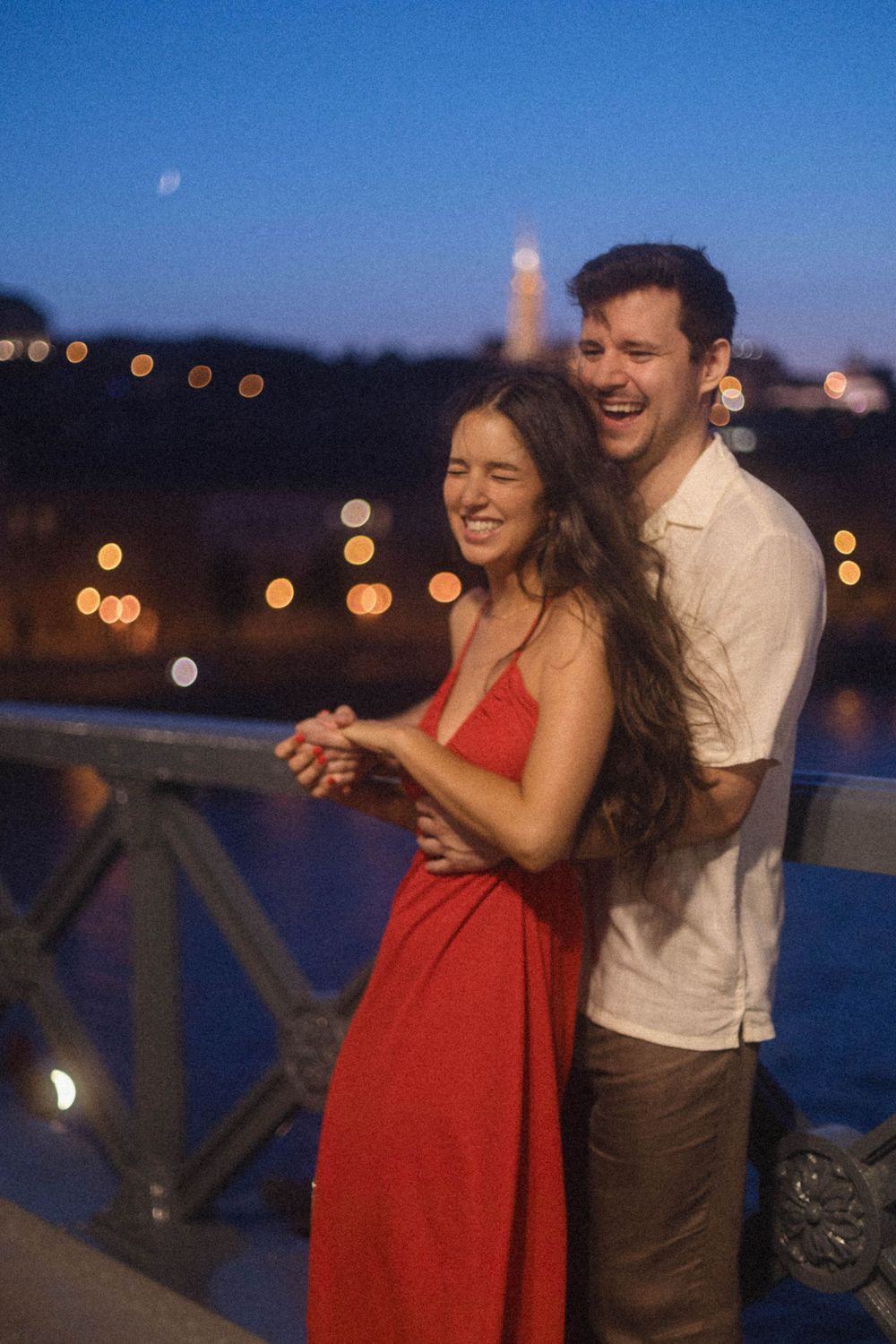 Laughing couple embracing on a bridge at night with bokeh city lights in Budapest.